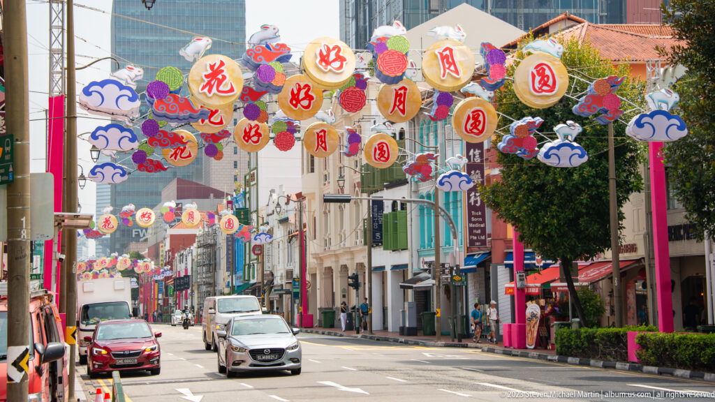 Singapore Chinatown street with overhead floral lanterns, cloud motifs, cars, shophouses, and modern buildings.