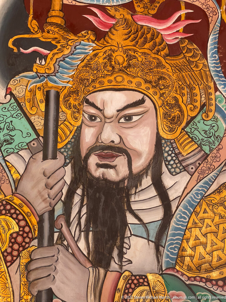 Close painted Chinese warrior guardian with black beard, gold helmet, patterned armor, and staff.