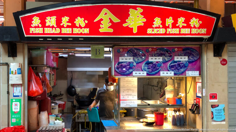 Sliced Fish Bee Hoon hawker stall with red sign, menu photographs, cook, and compact kitchen.