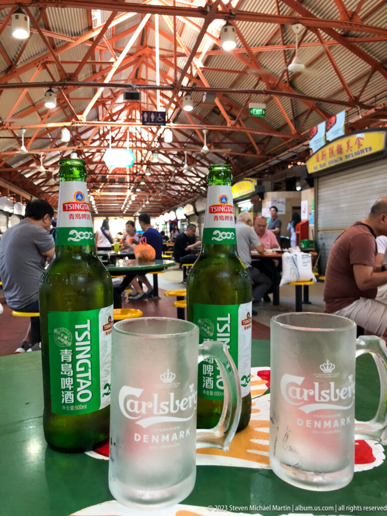 Two green Tsingtao bottles and frosted Carlsberg glasses on a table in a busy hawker centre.