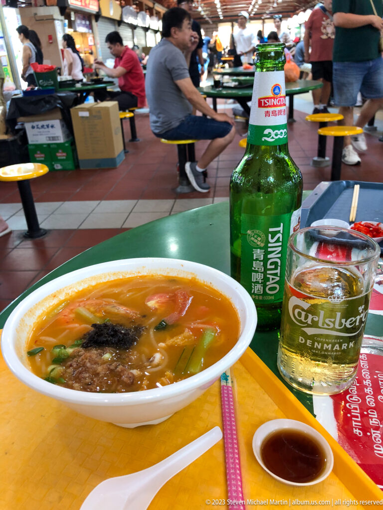Noodle soup on yellow tray with Tsingtao beer, glass, chopsticks, spoon, and dipping sauce.