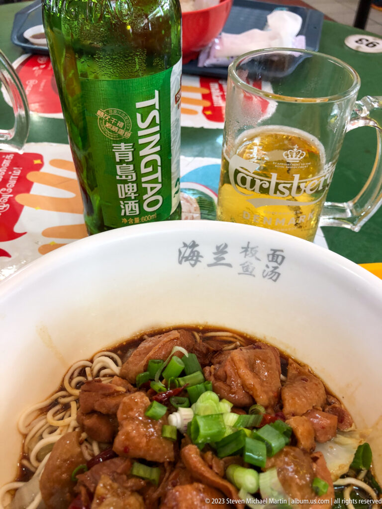 Dry noodles topped with braised meat and scallions beside Tsingtao beer and a glass.