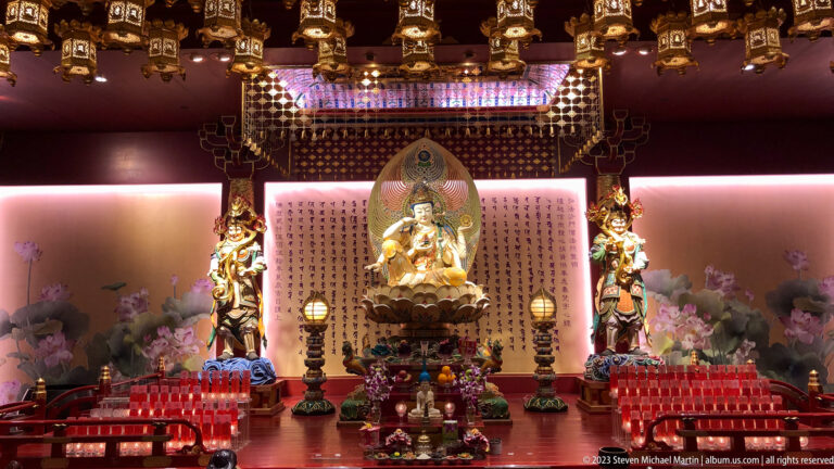 Seated bodhisattva on altar with attendants, red votive lights, flowers, lamps, and inscription panel.
