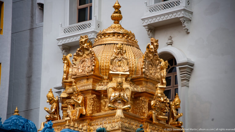 Highly gilded Hindu temple tower detail with small deity figures and ornate relief against a pale building.