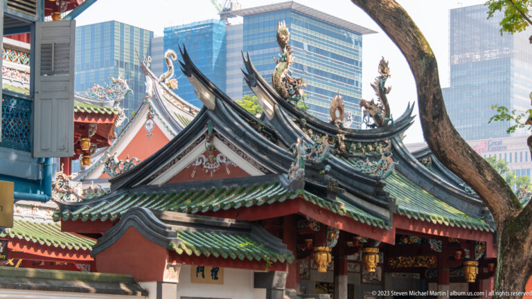 Side view of Chinese temple roofs with upturned black ridges, ceramic figures, tree, and modern towers.