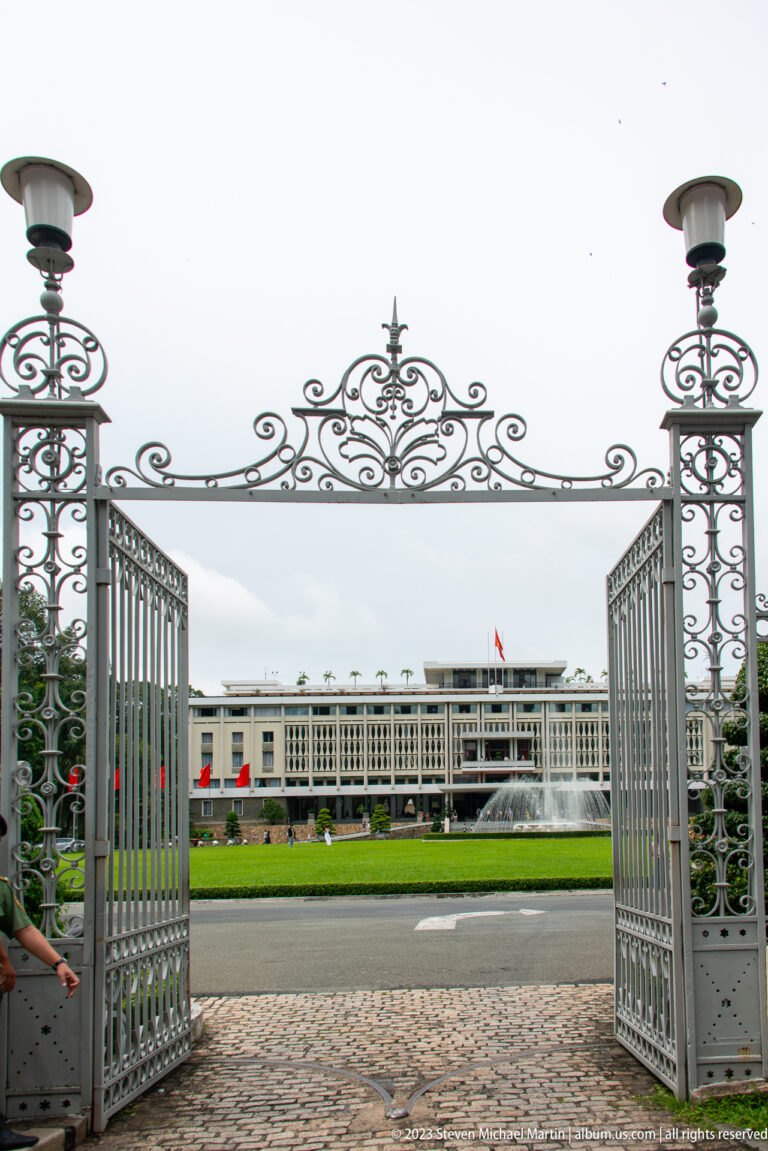 Open ornamental metal gates frame Independence Palace, its lawn, fountain, and Vietnamese flag in the distance.