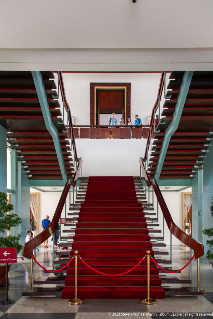 Broad red-carpeted staircase splitting into two flights beneath dark beams, with visitors on the upper gallery.