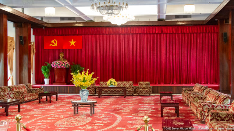 Formal red reception room with carved seating, floral displays, communist emblems, and a bust of Ho Chi Minh.