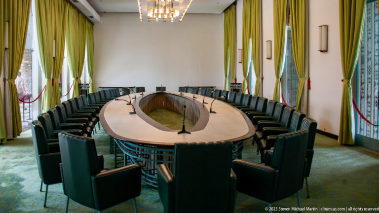 Oval conference table surrounded by green chairs and microphones in a curtained meeting room.