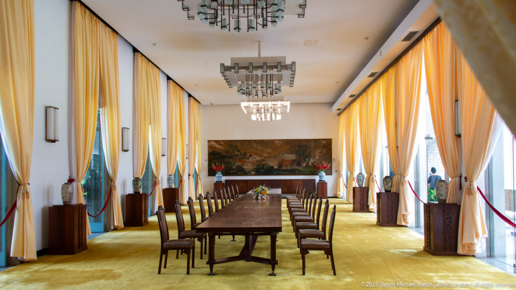 Long formal dining table with rows of chairs in a tall room with yellow curtains and display objects.