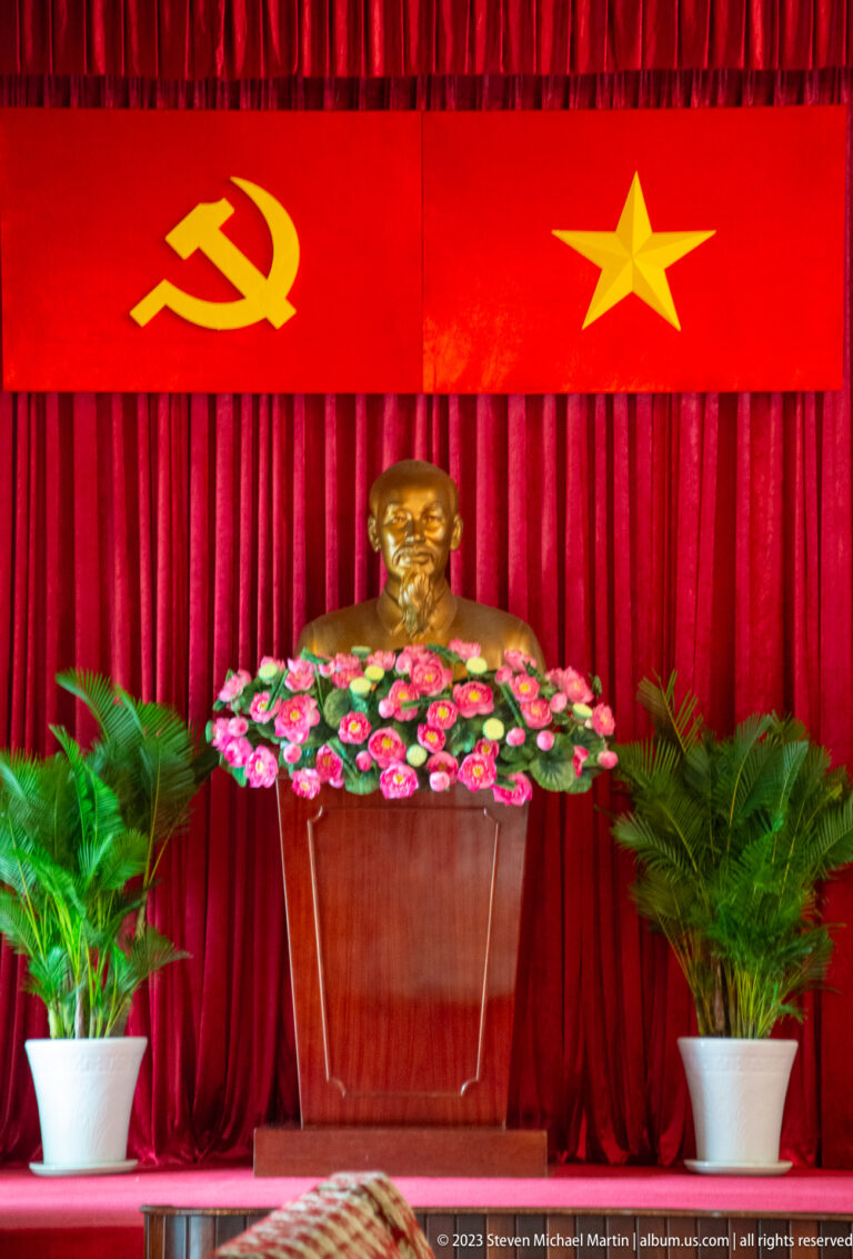 Gold-toned bust of Ho Chi Minh with pink flowers beneath a hammer-and-sickle and yellow star on red curtains.