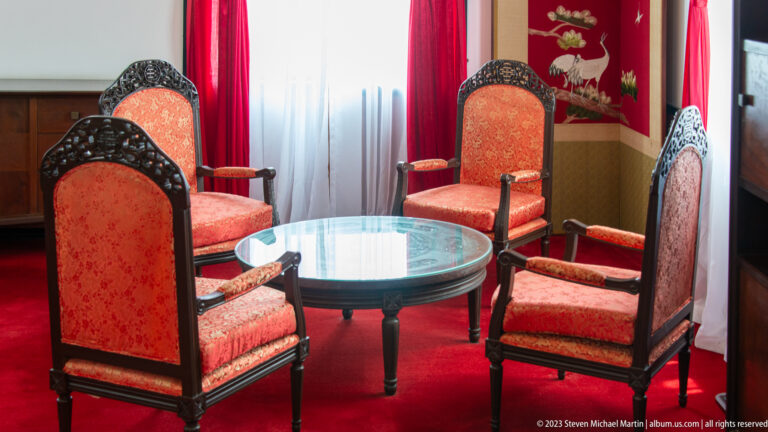 Four red upholstered wooden armchairs arranged around a circular glass-topped table on red carpet.
