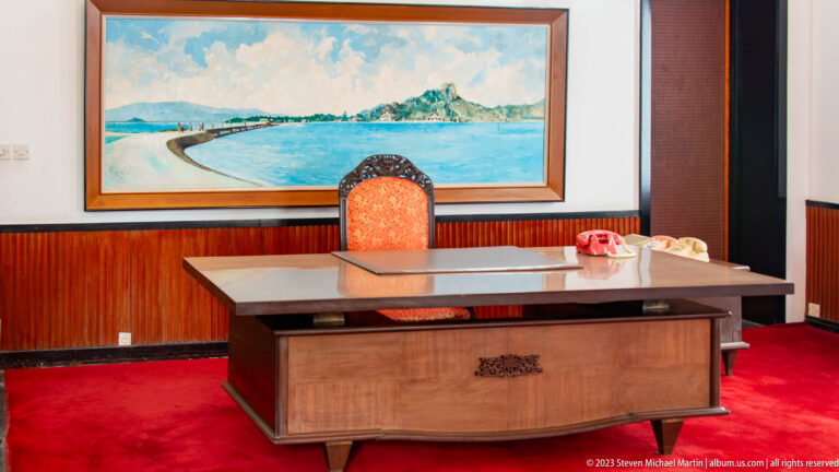 Wooden office desk with two rotary telephones beneath a coastal painting and an upholstered chair.