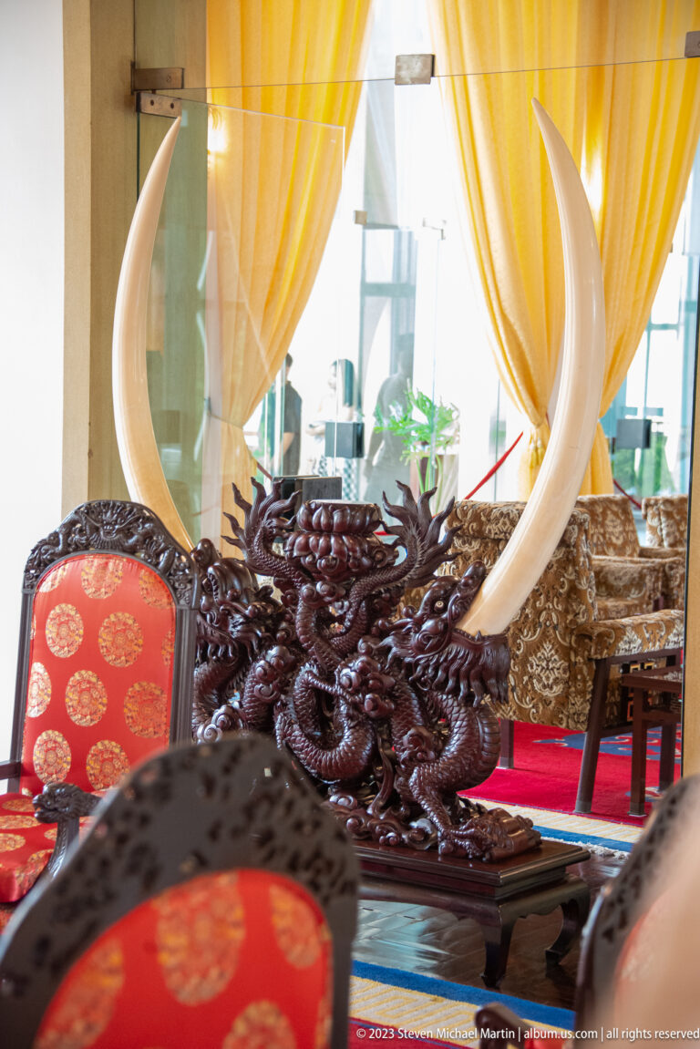 Two large curved tusks mounted on an ornate dark wooden stand among red upholstered chairs.
