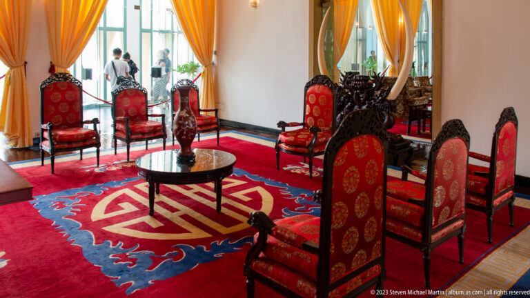 Formal reception room with red carved chairs, low tables, golden curtains, and a patterned dragon carpet.