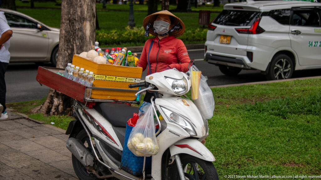 Masked street vendor beside a loaded motor scooter displaying bottled drinks, snacks, and hanging bags.