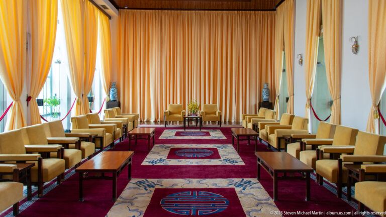 Long formal hall with paired armchairs, golden curtains, red runners, and circular symbols on patterned carpets.