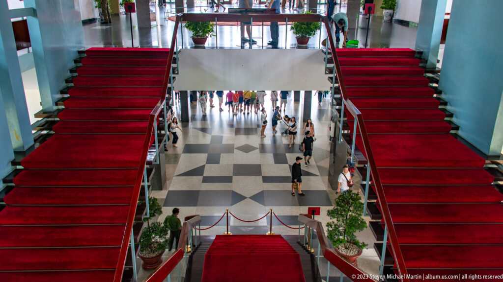 High view down two red-carpeted staircases toward a patterned lobby with visitors below.