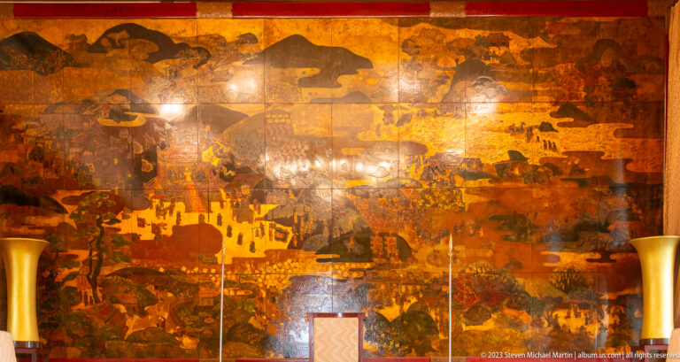 Close view of a large amber-and-brown lacquer mural showing figures, buildings, trees, and mountains.