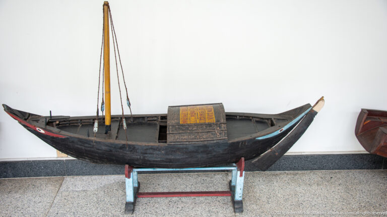 Long dark wooden boat model with upturned ends, central mast, and a small decorated cabin.