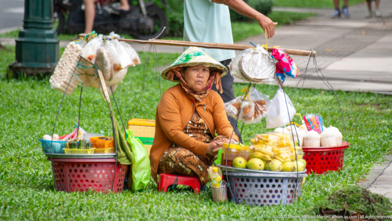 Seated vendor in a conical hat beside baskets of fruit and goods suspended from a wooden shoulder pole.