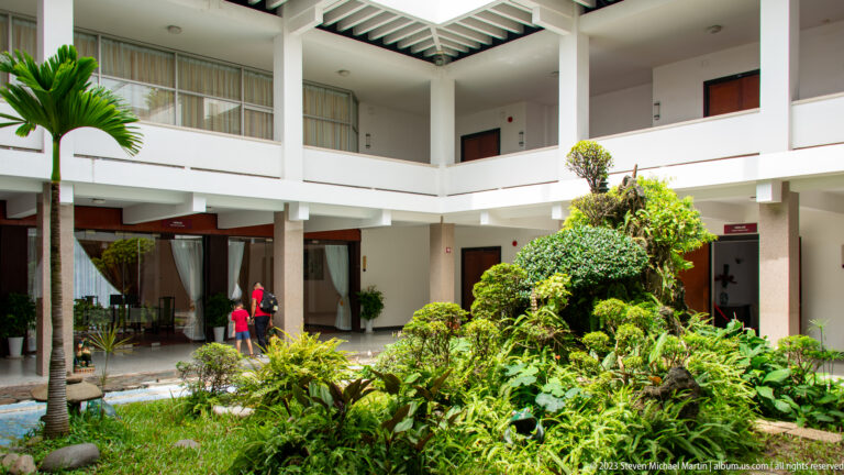 Interior courtyard with palms, ferns, clipped shrubs, white balconies, and open corridors.