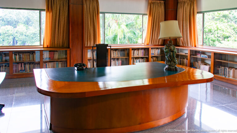 Wood-paneled library with a large curved desk, built-in bookshelves, corner windows, lamp, and telephone.