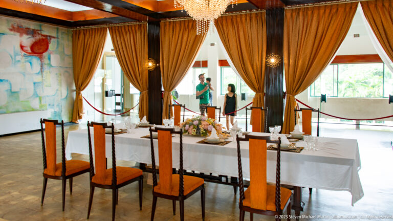 Formal dining table set with white linen, glassware, flowers, chandelier, and high-backed chairs.
