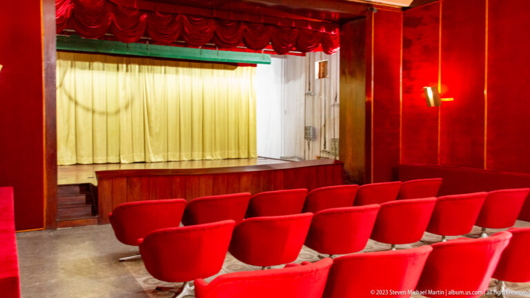 Small red auditorium with rows of upholstered seats facing a curtained stage.
