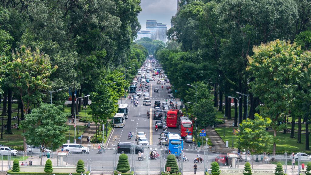 Elevated view down a busy tree-lined boulevard filled with motorbikes, buses, cars, and distant towers.