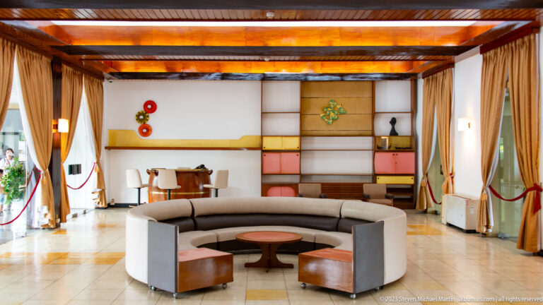 Bright lounge with a circular sectional sofa, low tables, geometric wall panels, and exposed ceiling beams.