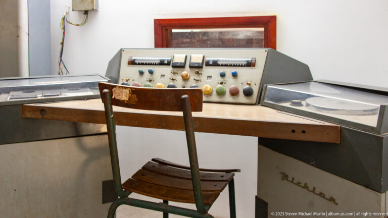 Vintage audio console with reels, switches, meters, knobs, and a wooden operator’s chair.