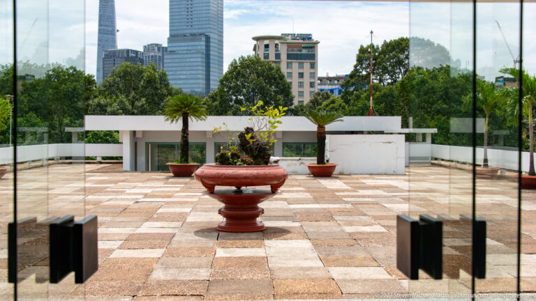 Tiled rooftop terrace with a large central planter, potted palms, low white walls, and city buildings beyond.