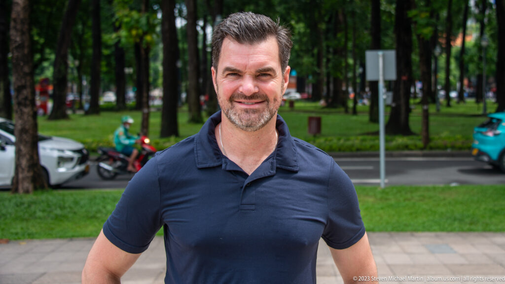 Smiling male visitor in a dark polo shirt standing on a tree-lined sidewalk near parked vehicles.
