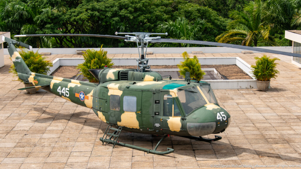 Camouflaged Bell UH-1 helicopter marked 445 displayed on the palace rooftop with its door open.