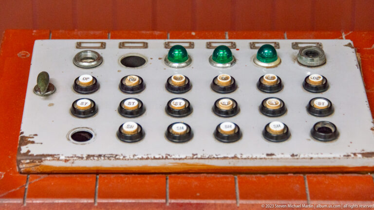 Metal control panel with rows of labeled buttons, green lights, switches, and sockets set into orange tile.