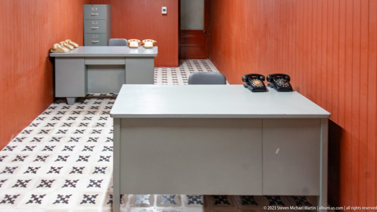 Narrow underground office with two metal desks, several rotary telephones, wood-paneled walls, and patterned tile floor.