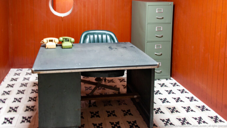 Metal desk with cream and green rotary telephones, green chair, and tall filing cabinet in a paneled room.