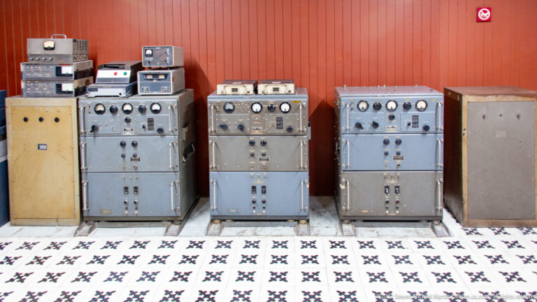 Row of large gray communications cabinets with dials, meters, switches, and stacked electronic units.