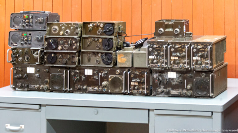 Stacked worn military radio sets with tuning dials, handles, speaker grilles, and metal cases on a table.
