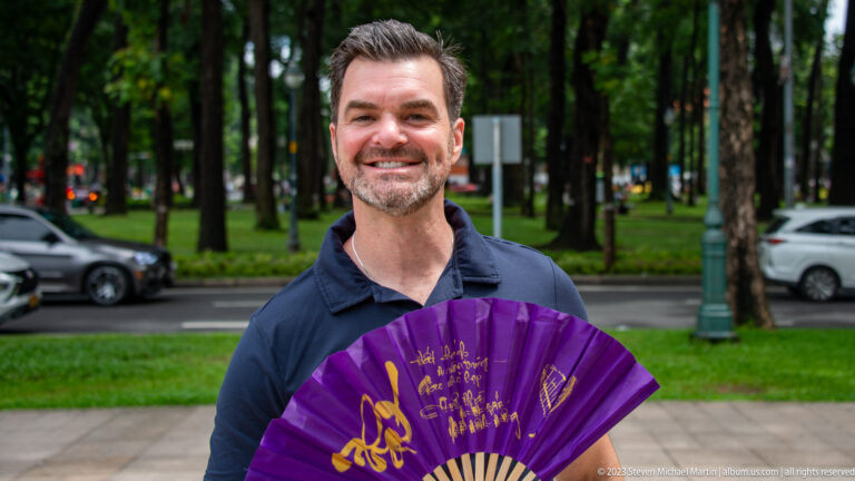 Smiling male visitor holding an open purple folding fan with gold decoration on a tree-lined sidewalk.