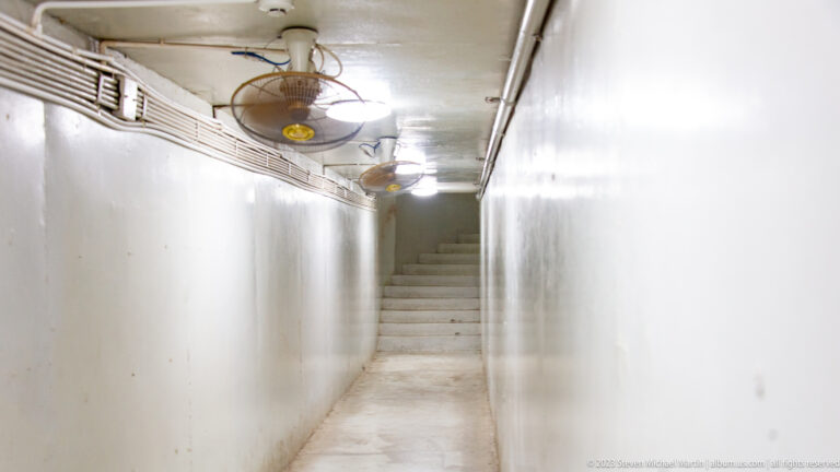 Narrow white underground corridor with exposed pipes, ceiling fans, conduits, and stairs at the far end.