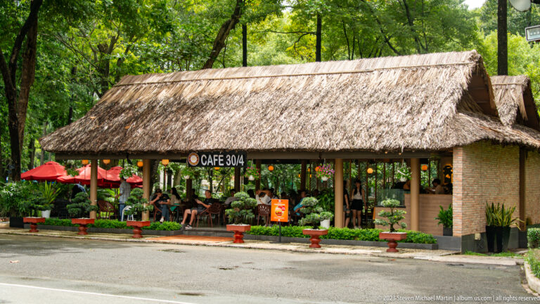Open-sided Café 30/4 pavilion with a thatched roof, shaded seating, and landscaped plants.