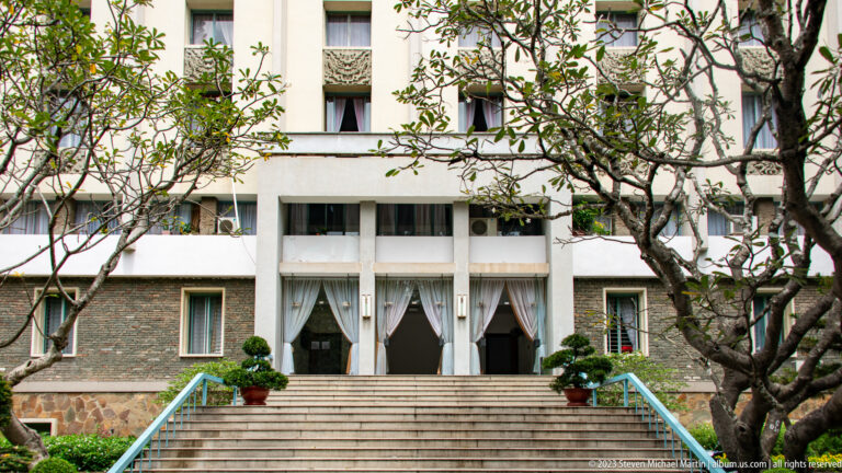 Broad steps with pale blue railings leading to a recessed doorway on a tree-framed palace façade.