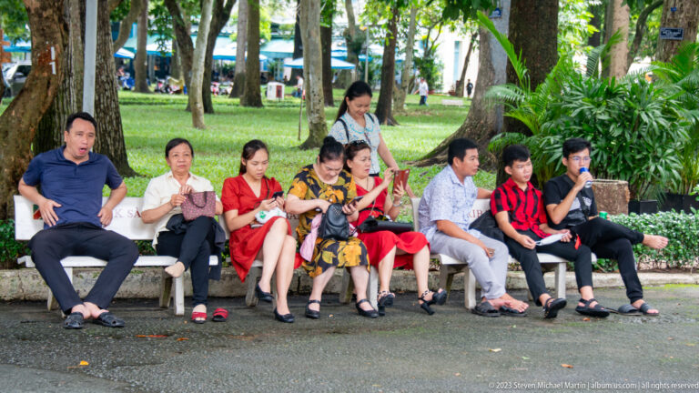 Group of visitors seated on a long bench beneath trees in the landscaped palace grounds.