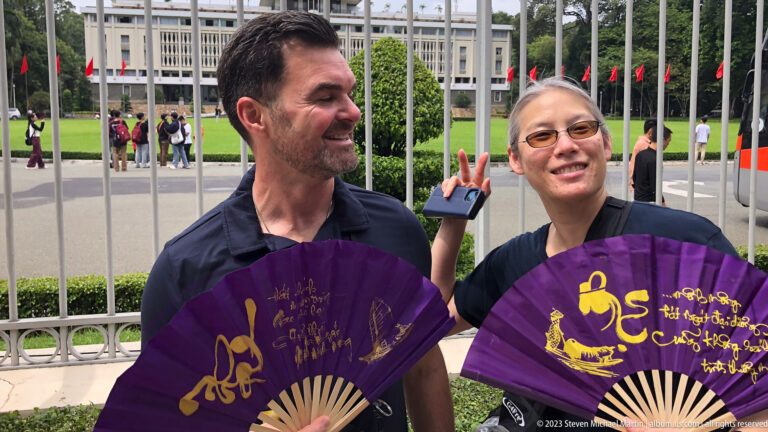 Two smiling visitors holding open purple-and-gold folding fans outside the palace fence.