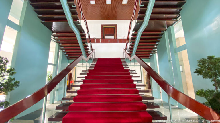 Low centered view up a red-carpeted staircase dividing into two flights beneath dark beams.