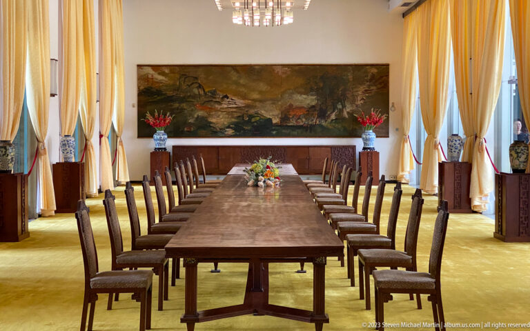 Long polished dining table with aligned chairs, gold curtains, chandeliers, flowers, and a wall painting.