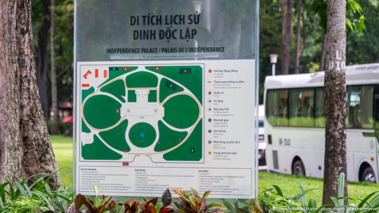 Bilingual Independence Palace site map mounted outdoors among trees, with a bus visible behind it.