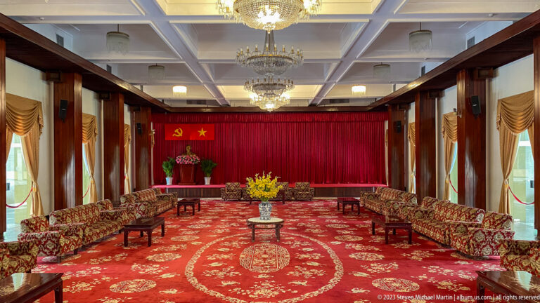 Wide ceremonial hall with patterned sofas, red curtains, chandeliers, communist emblems, and a Ho Chi Minh bust.